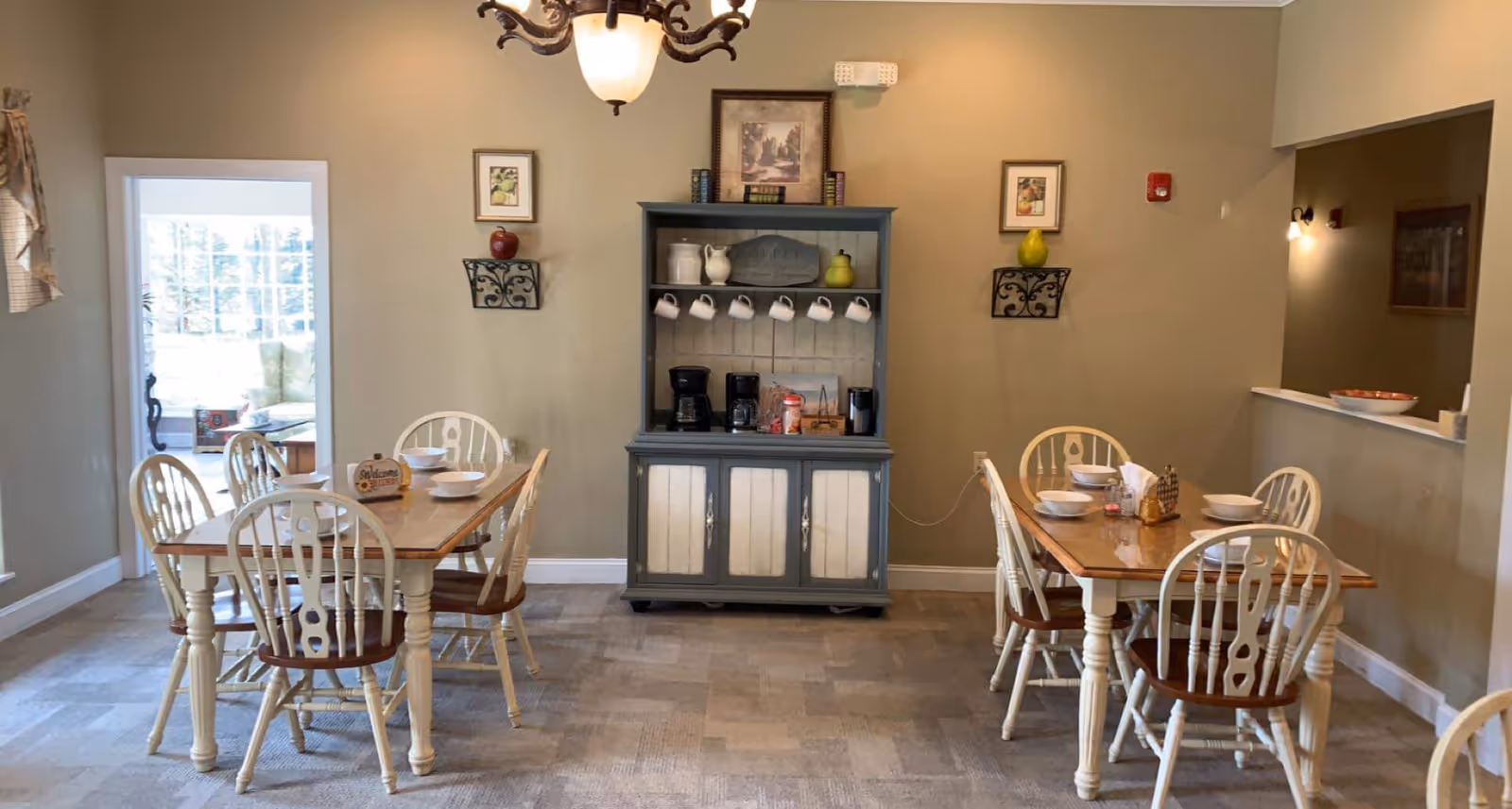 A cozy dining room with two wooden dining tables and chairs, a blue hutch with a coffee station against a beige wall, and soft overhead lighting.