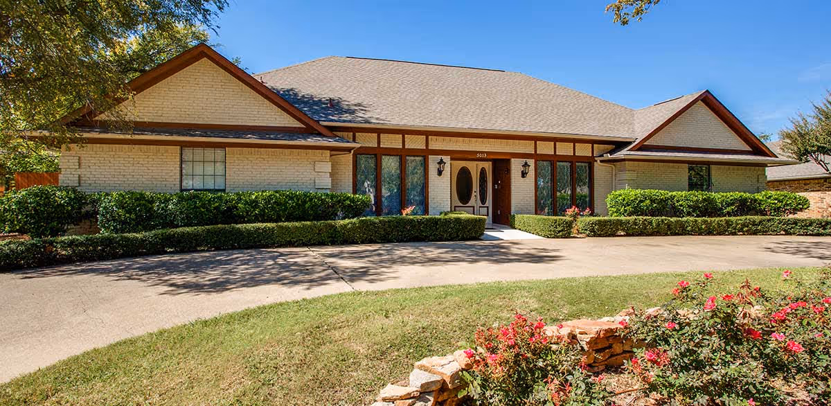 Single-story brick building with a large front entrance featuring double doors and multiple windows, surrounded by neatly trimmed bushes and a curved driveway, with a flower bed of red flowers in the foreground under a clear blue sky.