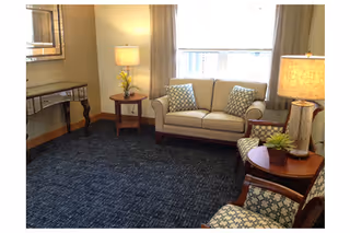 A cozy living room with a beige loveseat adorned with two patterned pillows, two patterned armchairs, a dark blue carpet, two wooden side tables each with a lamp, and a large window with curtains letting in natural light.