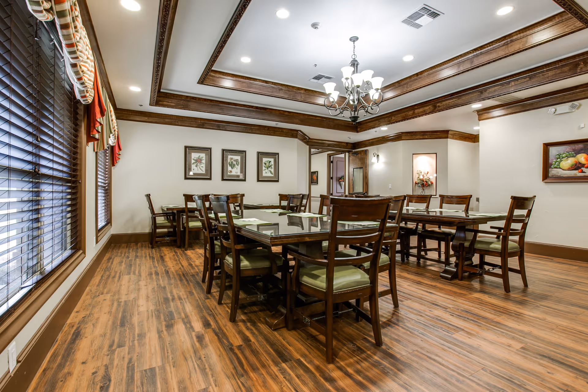 A well-lit dining room with wooden floors and multiple wooden tables and chairs arranged neatly. The room features large windows with blinds and valances, framed botanical artwork on the walls, and a decorative chandelier hanging from a tray ceiling with wooden trim.