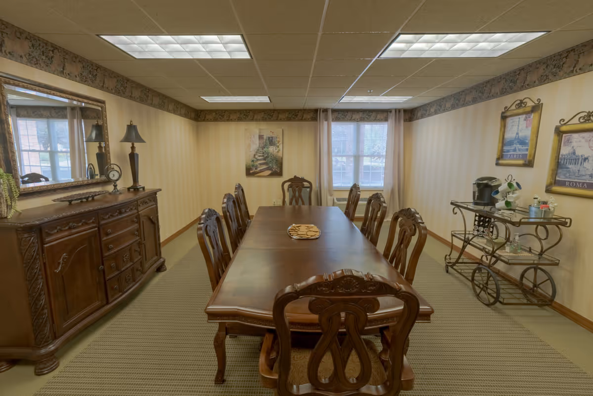 A formal dining room with a long wooden table surrounded by eight ornate wooden chairs. A plate of cookies is placed in the center of the table. The room has beige striped wallpaper with a floral border near the ceiling. On the left side, there is a wooden sideboard with a large mirror, a lamp, and a clock. On the right side, there is a metal serving cart with a coffee maker, cups, and other items. Two framed pictures hang on the right wall, and a window with sheer curtains is at the far end of the room.
