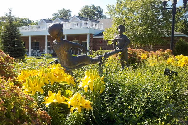 Bronze statue of two children playing amid yellow flowers in a garden in front of a brick building with a covered porch.