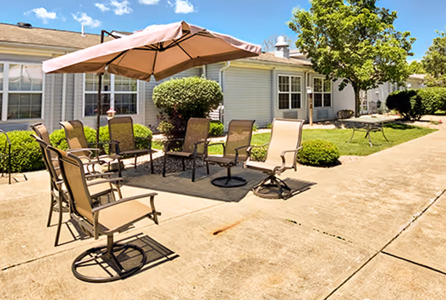 Outdoor patio area with several beige mesh chairs arranged around a large umbrella on a concrete surface. The patio is adjacent to a single-story building with white siding and multiple windows. Green bushes and a tree are visible along the building, under a clear blue sky.