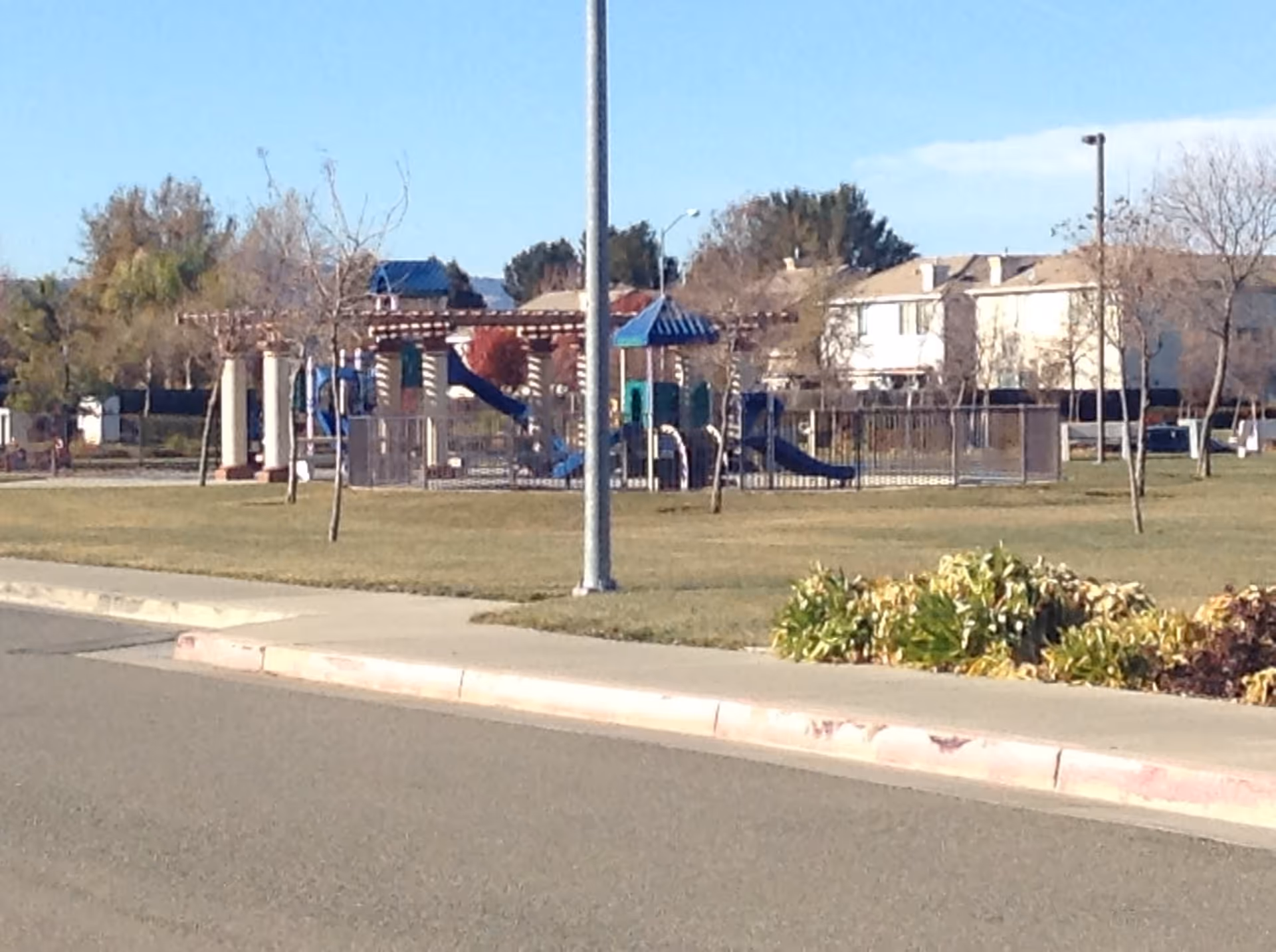 Outdoor playground area with slides and climbing structures surrounded by a grassy field, trees, and residential buildings in the background under a clear sky.