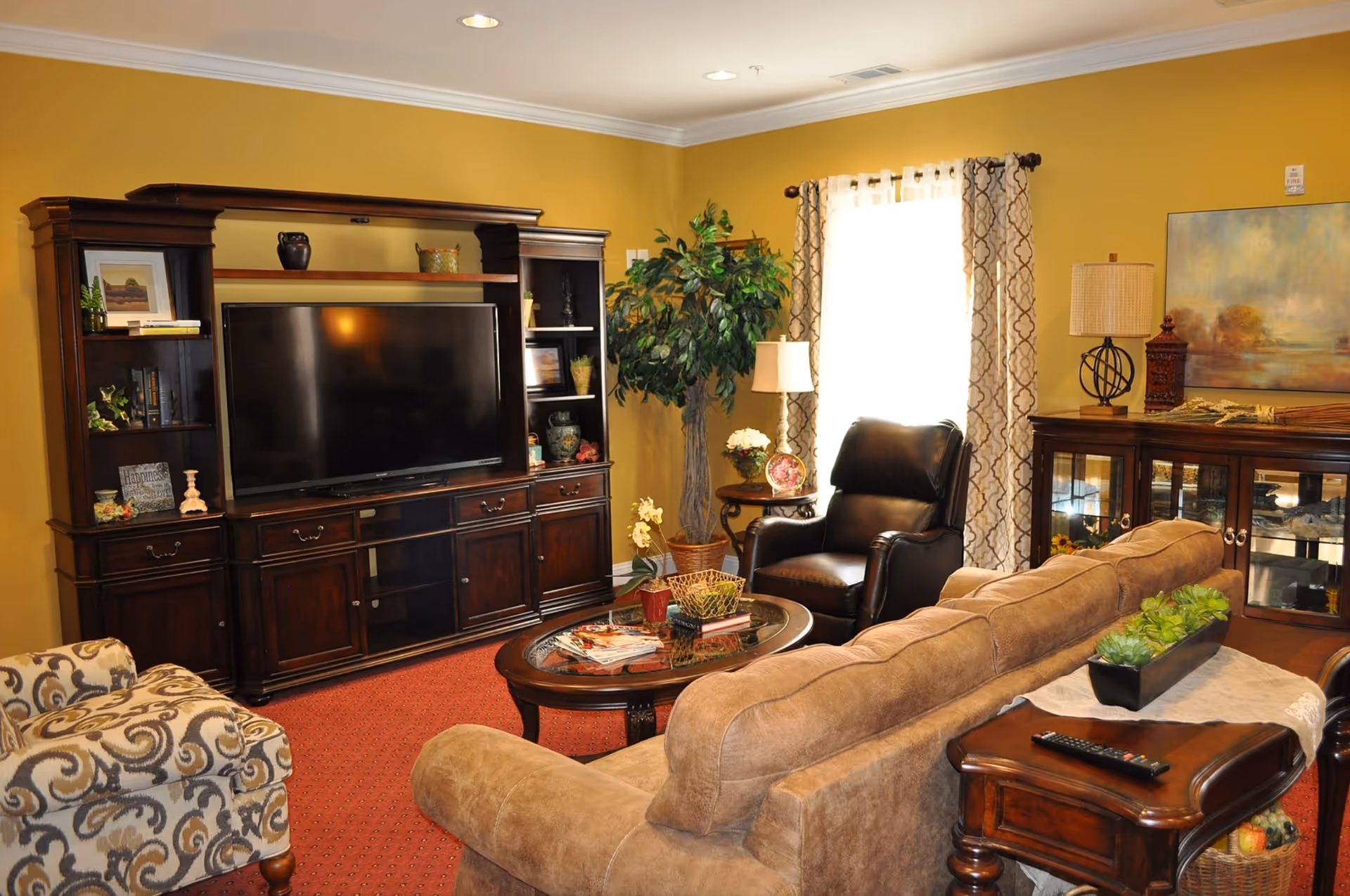 A cozy living room with a large flat-screen TV on a dark wooden entertainment center, a beige sofa, a patterned armchair, a black leather recliner, a wooden coffee table with magazines and plants, a side table with a lamp and flowers, and a window with patterned curtains letting in natural light.