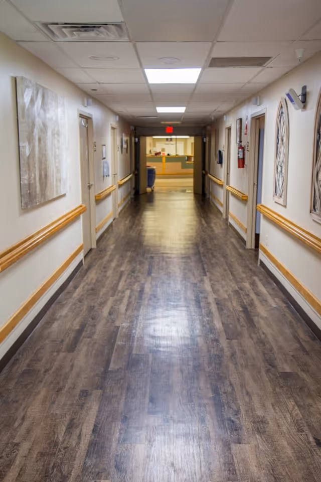 A long, clean hallway in a healthcare facility with wood-patterned flooring, beige walls, handrails on both sides, several closed doors, wall decorations, and a reception area visible at the far end under bright ceiling lights.