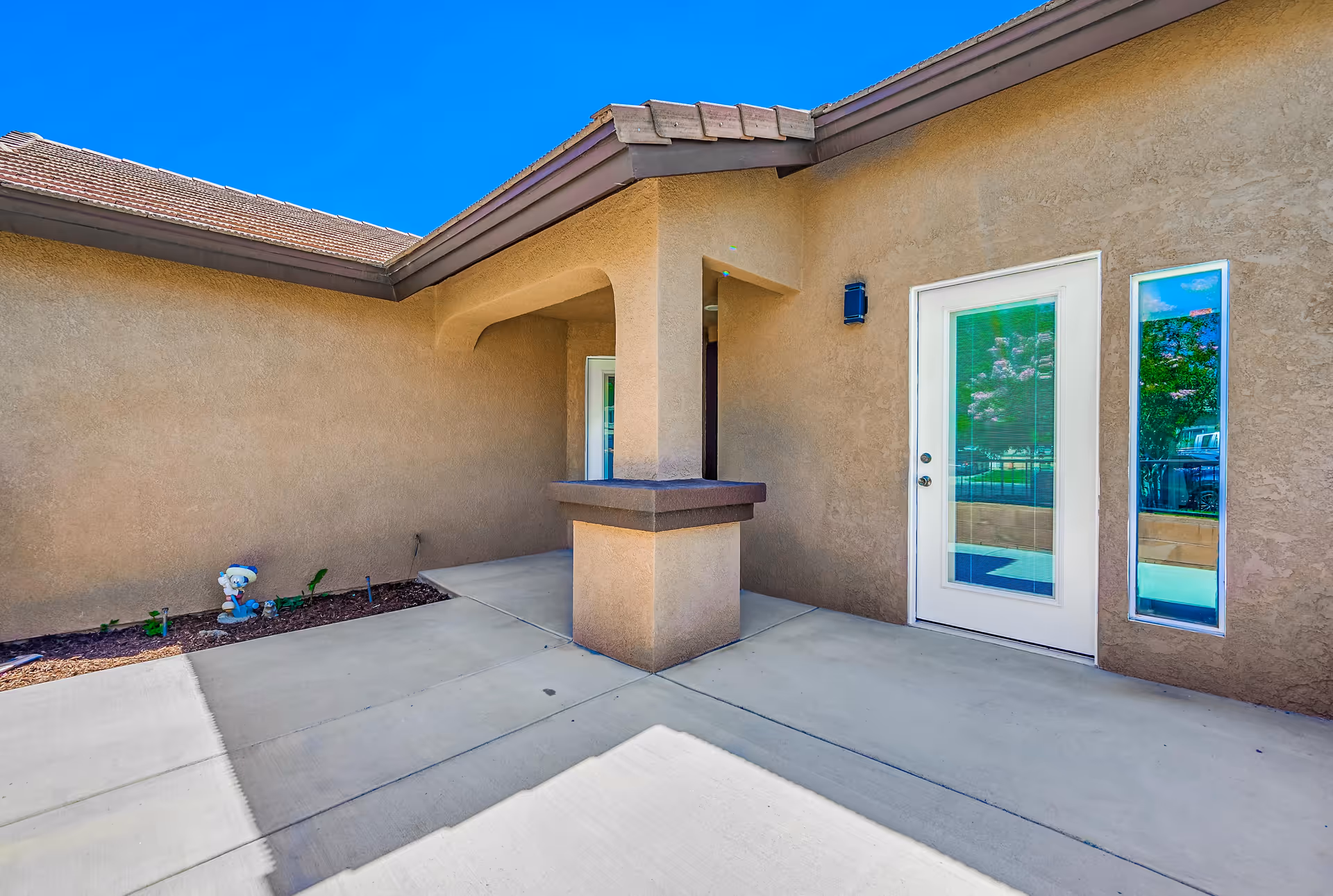 Covered stucco entry patio with a white glass door, narrow window, columned overhang, concrete walkway and a small planting bed with a garden figurine.