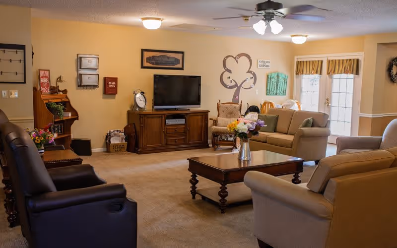 A cozy senior living community lounge with comfortable seating including armchairs and sofas arranged around a wooden coffee table with a flower vase. A flat-screen TV is mounted on a wooden cabinet against the wall. The room has warm beige walls decorated with framed pictures, a wall clock, and a decorative tree wall art. There is a ceiling fan with lights, and natural light enters through a glass door with striped valances.
