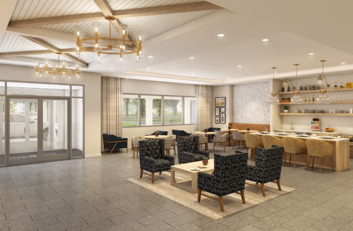 Spacious, well-lit senior living lobby/lounge with grouped armchairs around a coffee table, a dining counter with stools, chandeliers, and large windows by the entrance.