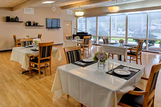A bright dining room with several tables covered in white tablecloths, each set with plates, glasses, and floral centerpieces. Wooden chairs surround the tables, and large windows on one side let in natural light. A piano and a wall-mounted TV are visible in the background.