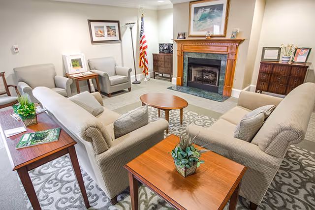 A cozy living room with beige upholstered sofas and armchairs arranged around a wooden coffee table. The room features a fireplace with a wooden mantle, a framed painting above it, and an American flag in the corner. There are wooden side tables with plants and magazines, a patterned carpet, and soft lighting.