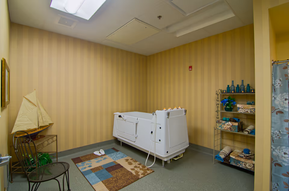 Walk-in bathtub in a softly lit assisted-living bathing room with a decorative rug, shelving of towels and bottles, a small chair and a model sailboat.