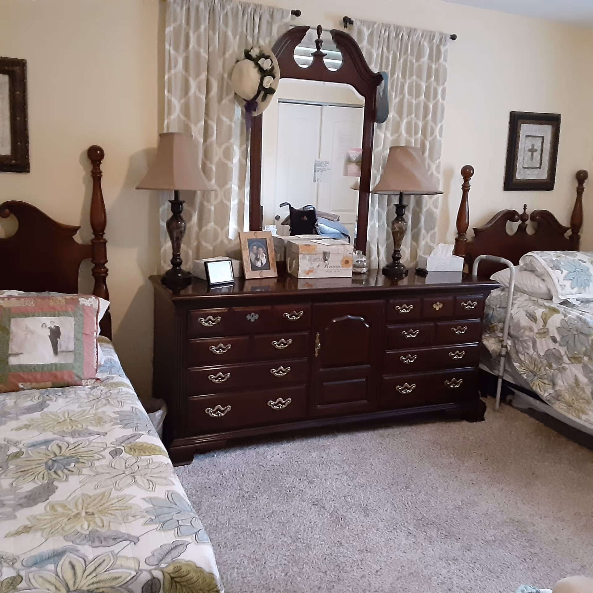 A bedroom with two twin beds on either side of a dark wooden dresser with a large mirror. Each bed has floral patterned bedding and a pillow with a photo print. The dresser has two matching lamps, framed photos, a decorative box, and a tissue box. Beige curtains hang behind the dresser, and the room has light-colored carpet.