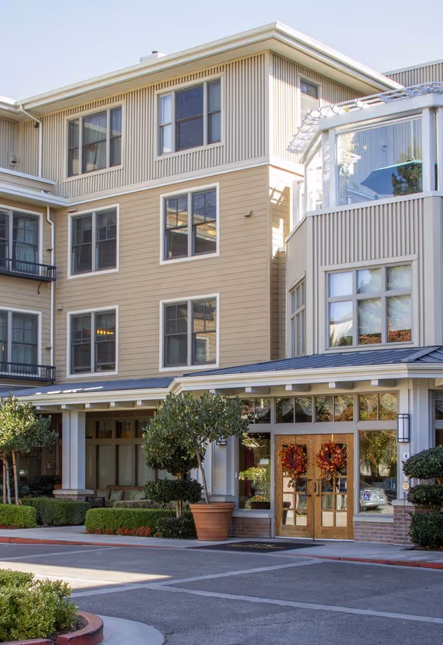 Exterior view of a multi-story senior living facility building with beige siding and large windows. The entrance features double wooden doors decorated with autumn wreaths, flanked by potted plants and small trees. The area is well-maintained with shrubs and a paved driveway in front.