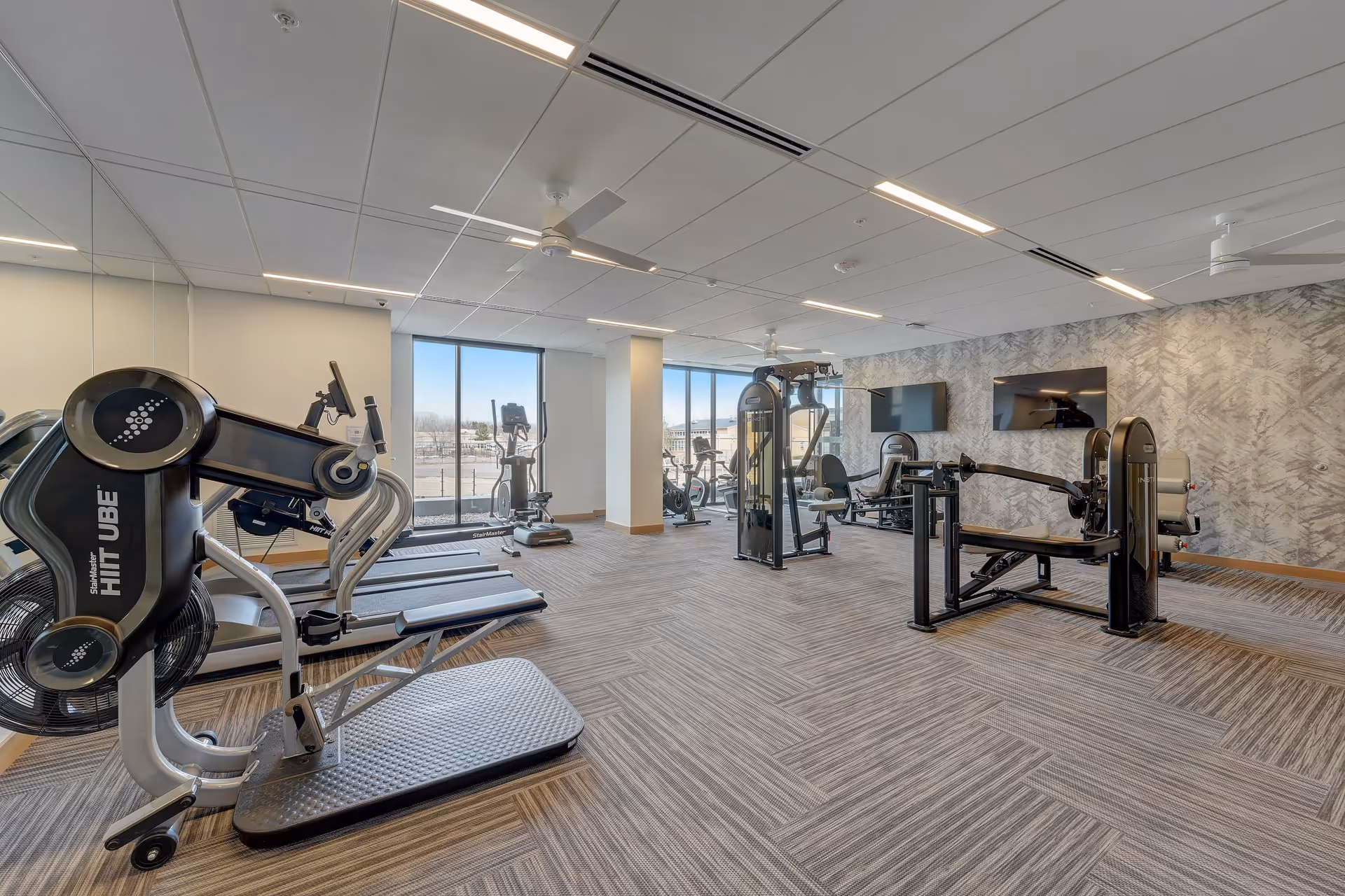 A modern fitness room with various exercise equipment including treadmills, stationary bikes, and weight machines. The room has large windows allowing natural light to enter, a patterned carpet floor, and two wall-mounted flat-screen TVs. The walls feature a textured design and the ceiling has recessed lighting and ceiling fans.