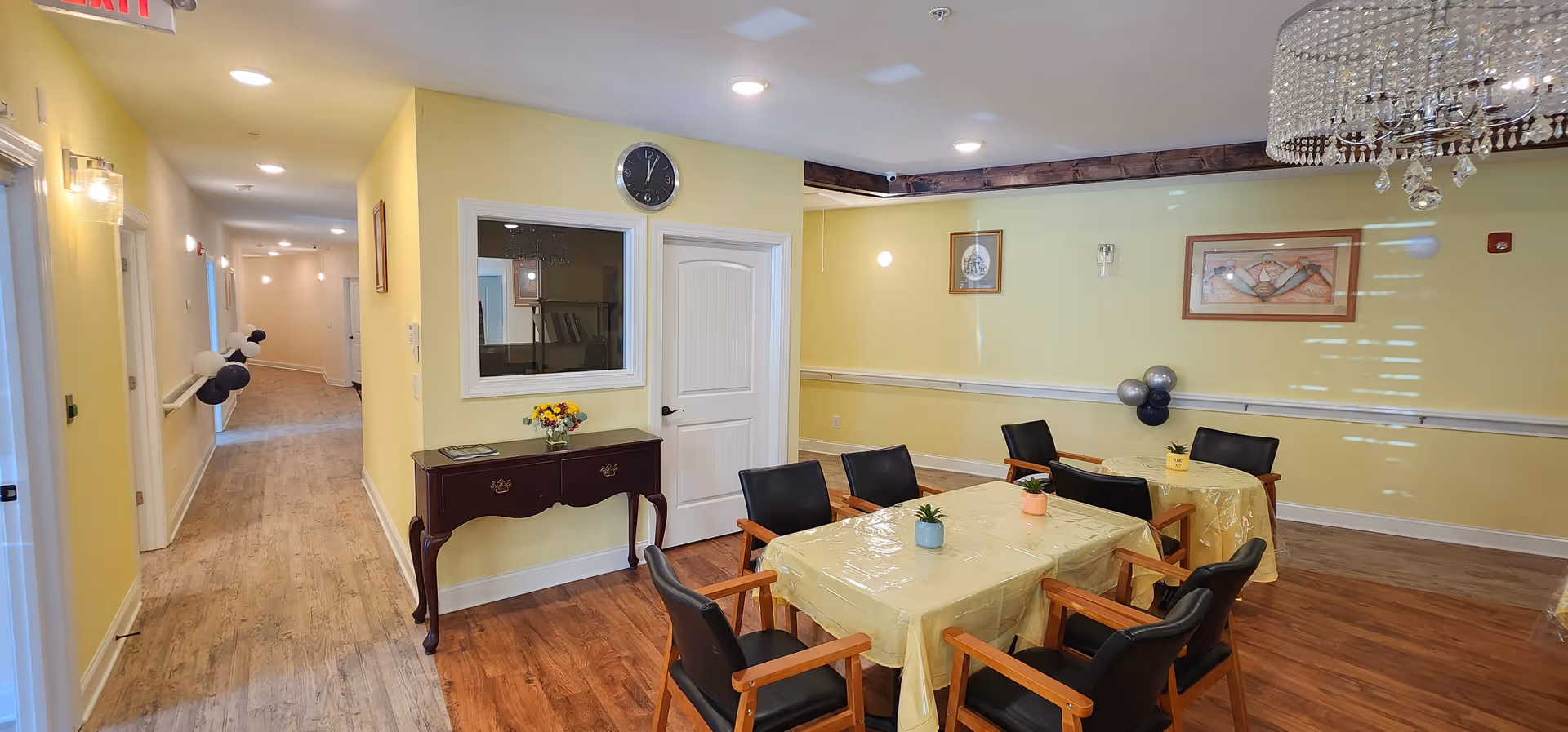 Interior view of a senior care facility room with yellow walls and wooden flooring. There are two tables covered with yellow tablecloths, each surrounded by black chairs with wooden armrests. A small plant is placed on each table. A chandelier hangs from the ceiling. On the left side, there is a hallway with light-colored flooring and wall-mounted lights. A small wooden console table with a flower vase is positioned against the wall near a door and a window. Wall decorations include framed pictures and balloons.