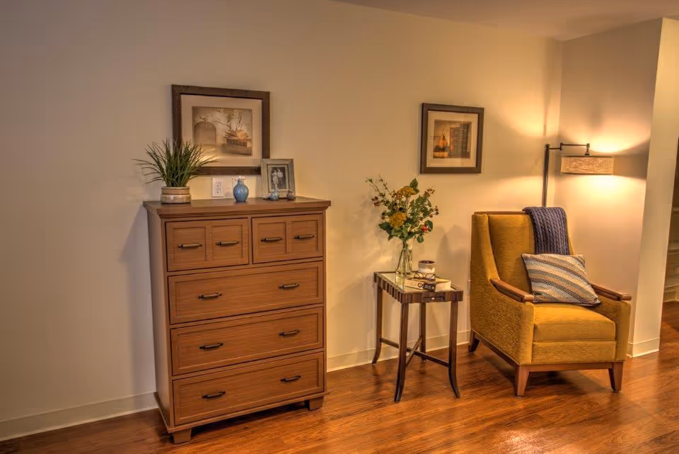 Cozy living area with a wooden chest of drawers, a small side table with flowers, and a mustard-yellow armchair under a floor lamp.