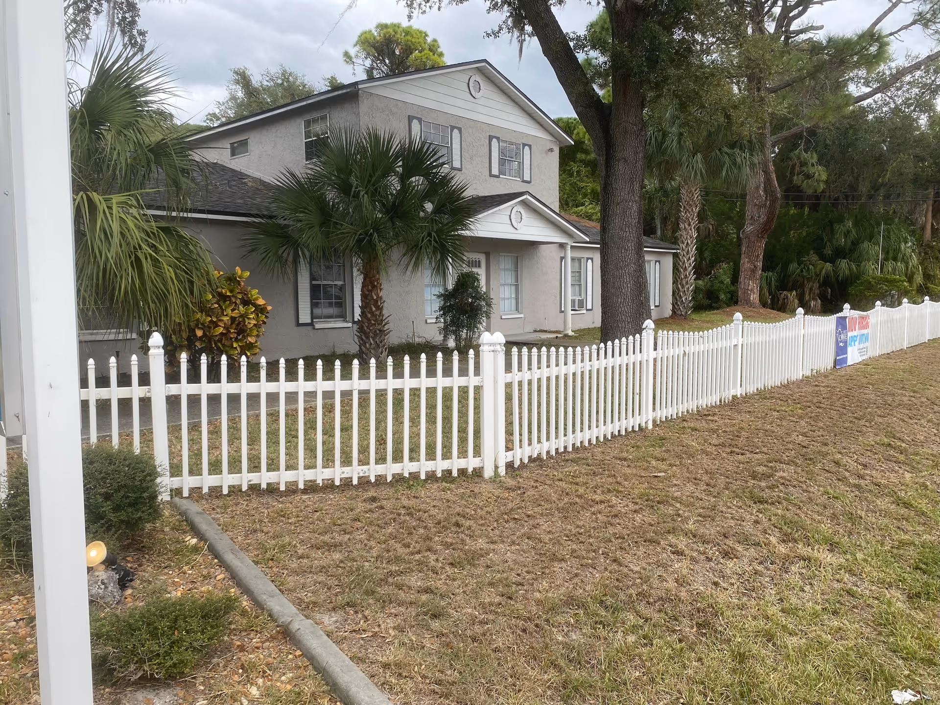 Two-story light-gray house with a white picket fence, palm trees, and a grassy front yard.