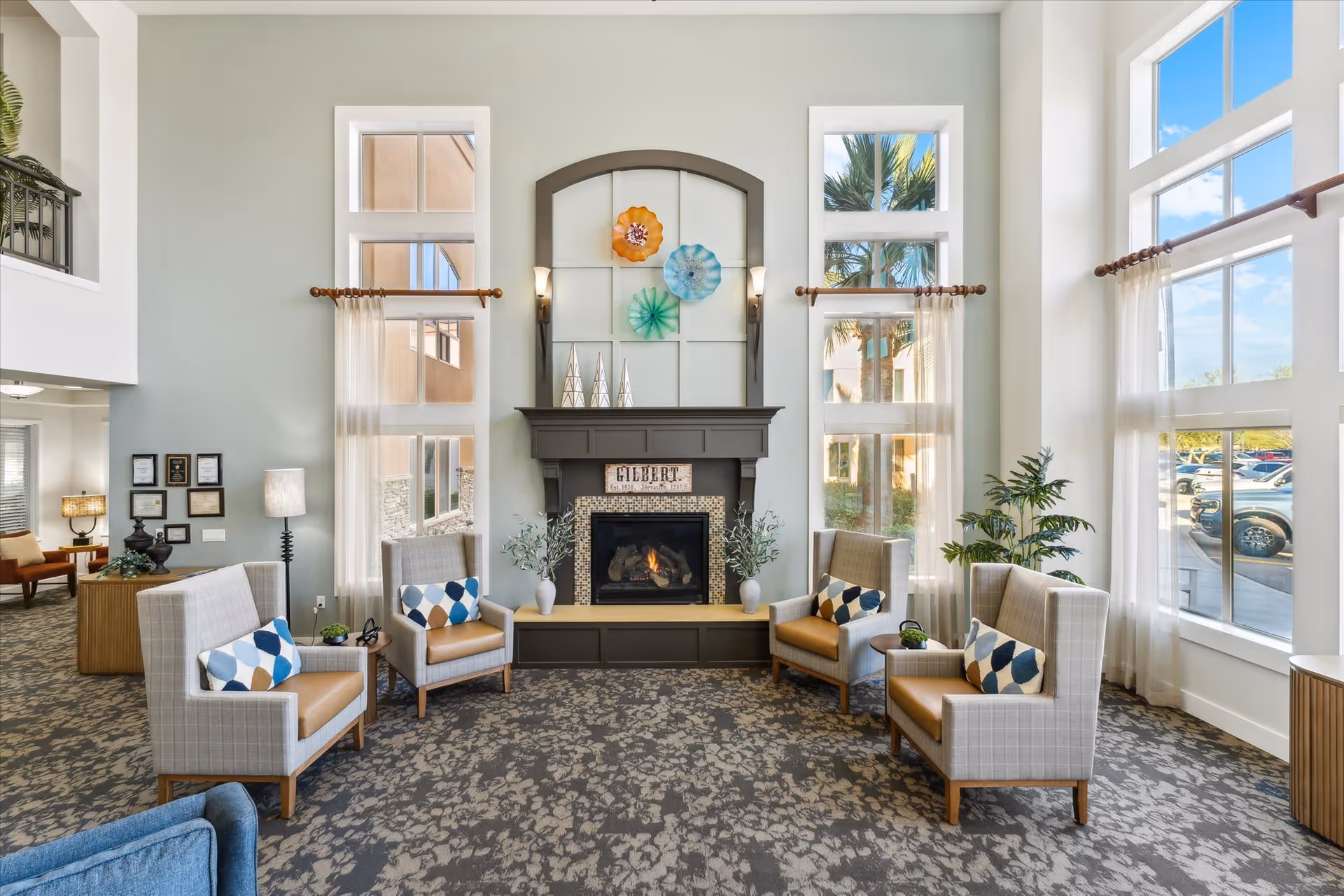 A bright and spacious living room area with large windows letting in natural light. The room features a fireplace with decorative art above it, surrounded by four cushioned armchairs with patterned pillows. There are plants on either side of the fireplace and a patterned carpet covering the floor. Outside the windows, cars and palm trees are visible.