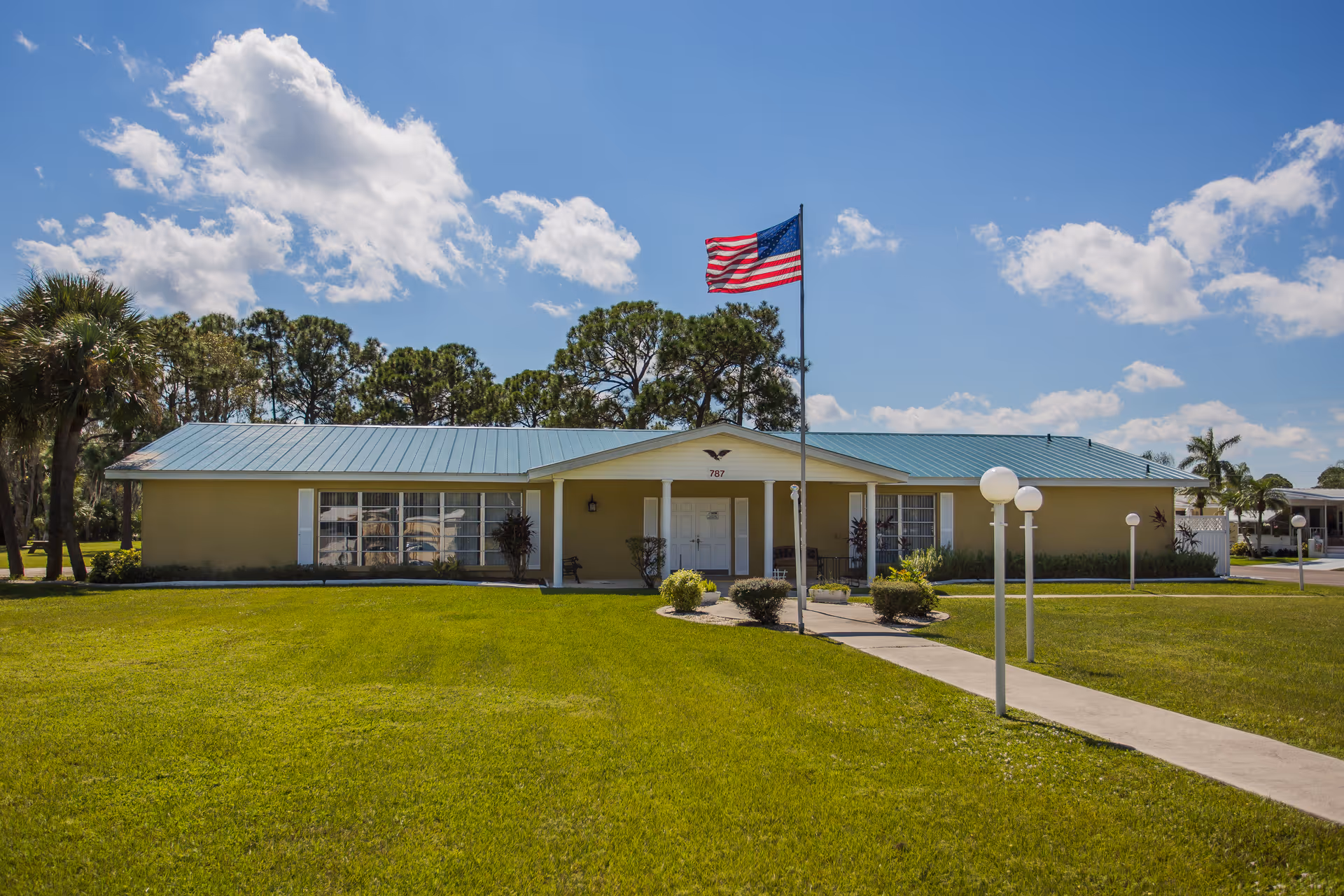 Single-story building with a light blue metal roof and beige walls, surrounded by a well-maintained green lawn. An American flag flies on a flagpole in front of the building, which has a covered entrance with double doors. The sky is blue with scattered clouds, and there are trees in the background.