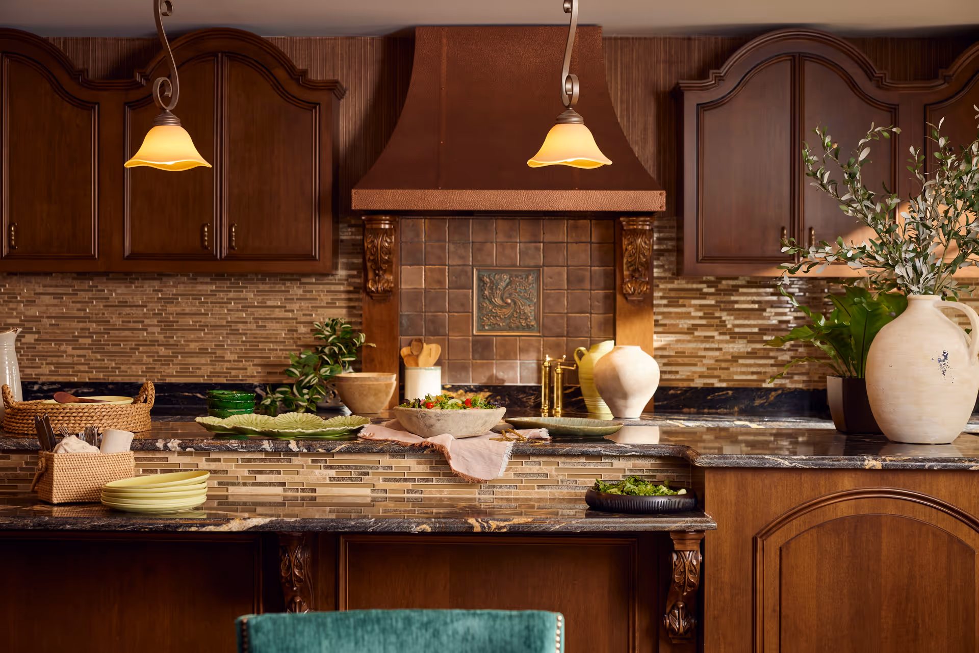 A warm, inviting kitchen with dark wooden cabinets, a tiled backsplash, and a large countertop adorned with various bowls, plates, a basket, and decorative plants. Two pendant lights hang from the ceiling, illuminating the space.