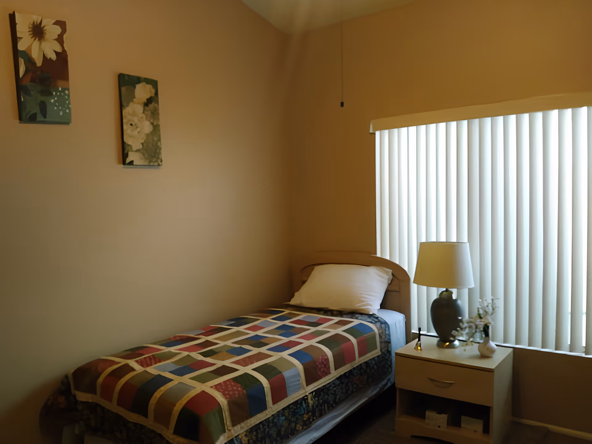 A small bedroom with a single bed covered by a colorful patchwork quilt. There is a white pillow on the bed. Next to the bed is a wooden nightstand with a lamp, a small vase with flowers, and a small decorative item. The window behind the nightstand has vertical blinds partially closed. Two floral paintings hang on the beige wall above the bed.