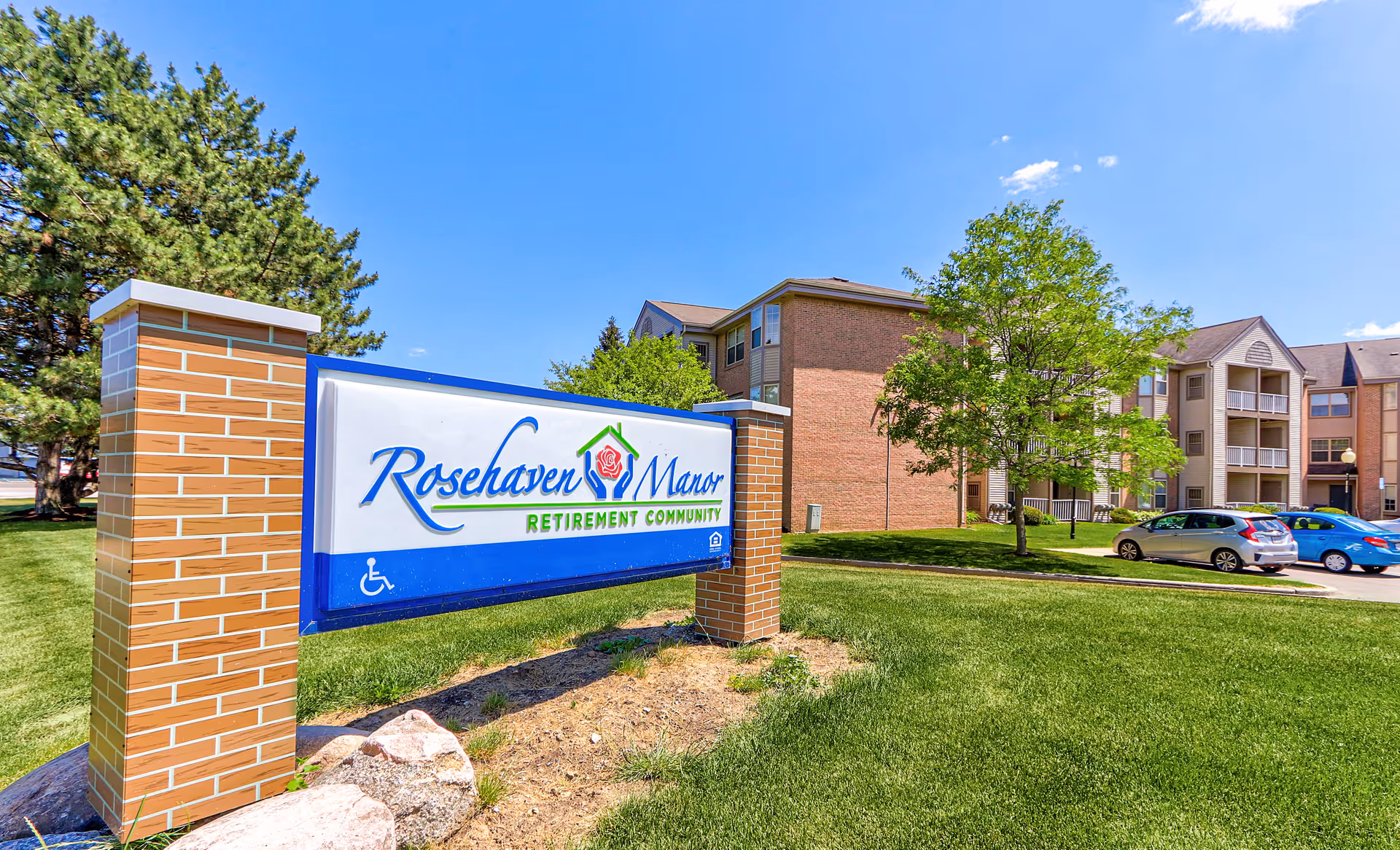 Outdoor view of Rosehaven Manor retirement community sign with brick pillars, green grass, trees, parked cars, and multi-story residential buildings under a clear blue sky.