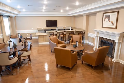 A cozy common area with wooden floors featuring a seating arrangement of four brown leather armchairs around a round wooden table near a white fireplace with a framed picture above it. In the background, there is a counter with bar stools and a mounted flat-screen TV. Additional round tables with chairs are placed near large windows with curtains.