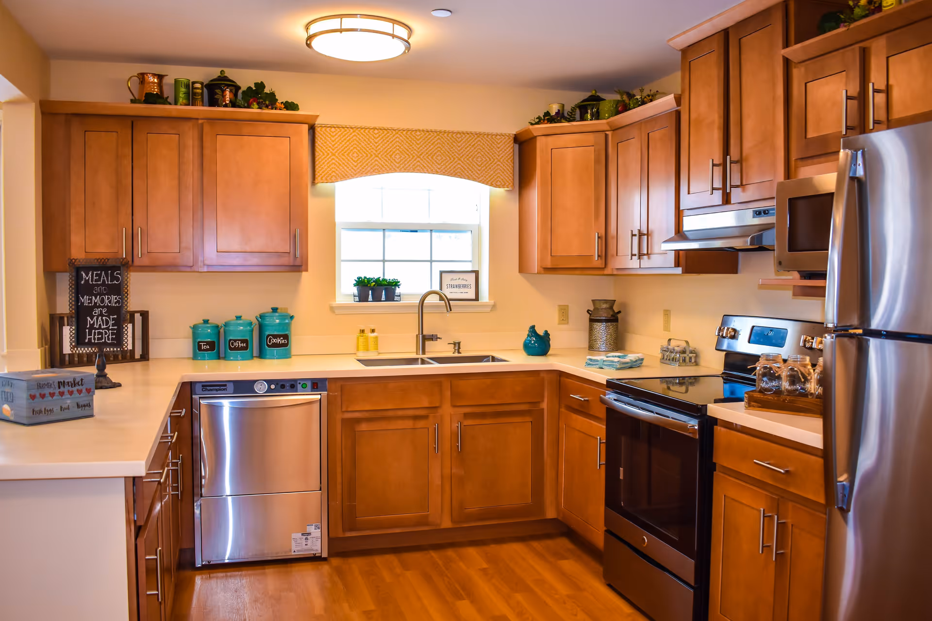 A modern kitchen with wooden cabinets, stainless steel appliances including a dishwasher, oven, microwave, and refrigerator. The kitchen has a white countertop with teal canisters labeled Tea, Coffee, and Cookies, a double sink under a window with a yellow valance, and decorative items on top of the cabinets.
