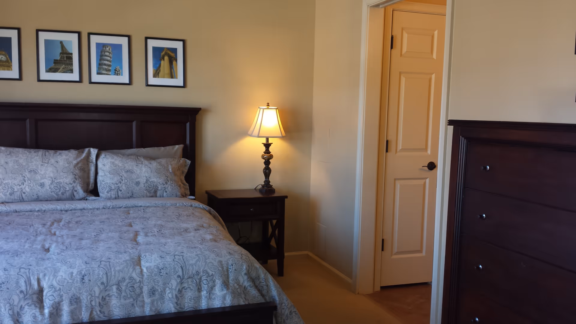 A bedroom with a large bed featuring a patterned comforter and pillows, a wooden headboard, a nightstand with a decorative lamp, a partially open door, and a wooden dresser. Four framed pictures hang on the wall above the bed.