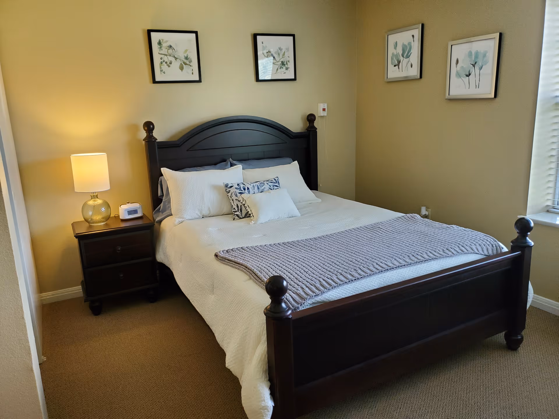 A neatly made bed with a dark wooden frame and headboard in a bedroom. The bed has white bedding with multiple pillows and a folded gray knitted blanket at the foot. A nightstand with a lamp and a small clock is beside the bed. Four framed botanical prints hang on the beige walls, and a window with blinds is visible on the right side.