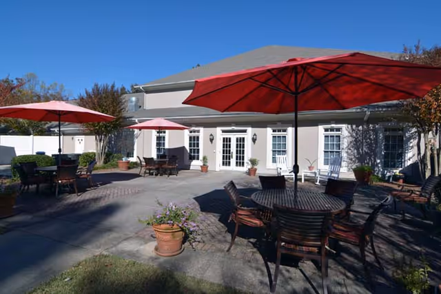 Outdoor patio area at Charter Senior Living of Buford with multiple round tables and chairs under large red umbrellas, potted plants, and a building with white doors and windows in the background under a clear blue sky.