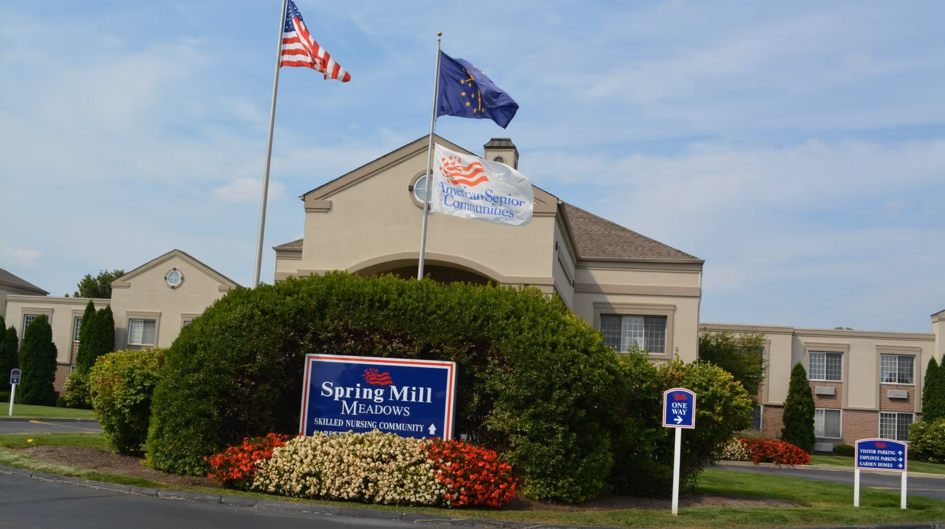 Front entrance of Spring Mill Meadows skilled nursing community with flags, a sign, bushes, and flowers.