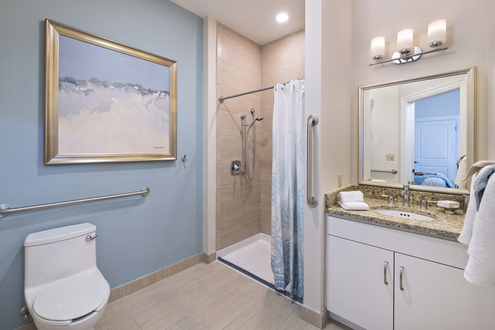 A clean, modern bathroom featuring a white toilet, a walk-in shower with a curtain, and a vanity with a granite countertop, sink, and mirror. The walls are painted light blue and beige, with a framed abstract painting above the toilet. Towels are neatly placed on the countertop and hanging on a rack.