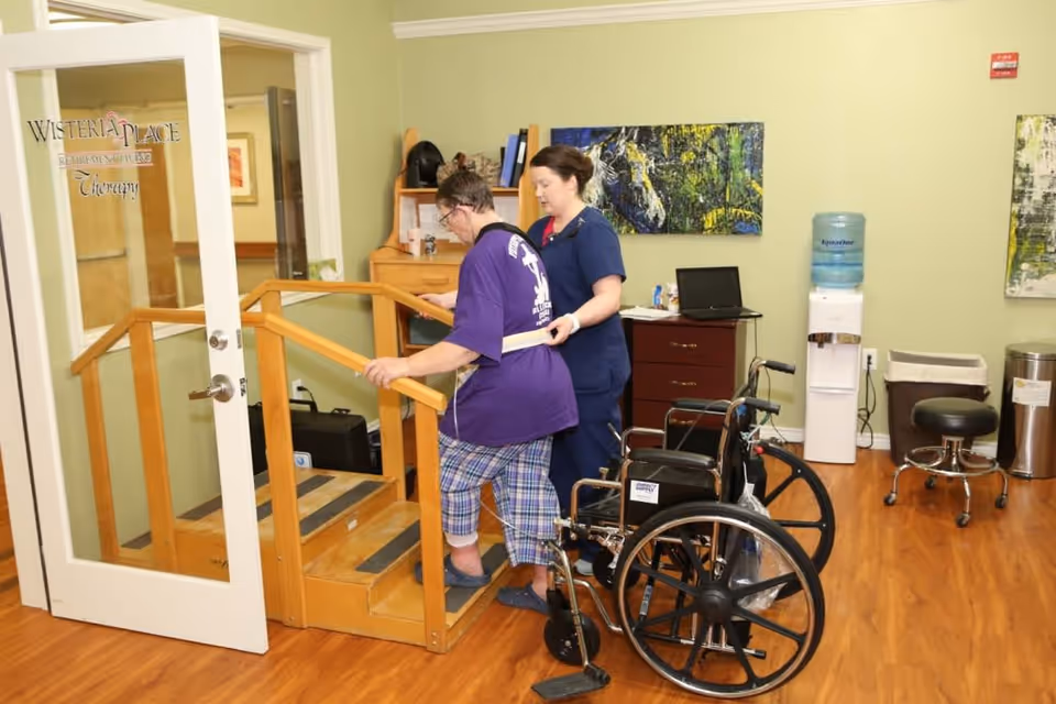 A caregiver assists an elderly woman using a gait belt as she practices walking up wooden steps with handrails inside a therapy room. A wheelchair is nearby, and the room has wooden flooring, a water cooler, a small cabinet with a laptop, and colorful artwork on the walls.