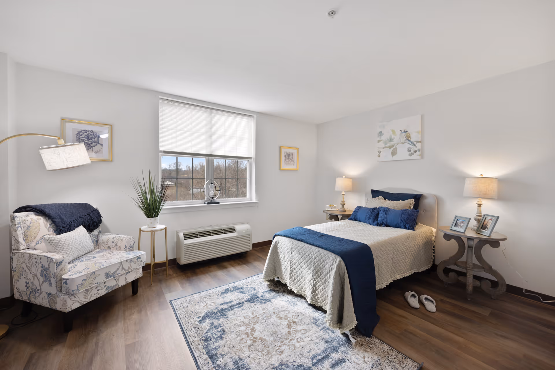A bright and cozy bedroom with a large window letting in natural light. The room features a neatly made bed with beige and navy blue bedding, two bedside tables with lamps, framed artwork on the walls, a patterned armchair with a throw blanket and pillow, a small side table with a plant, and a decorative rug on the wooden floor. Slippers are placed beside the bed.