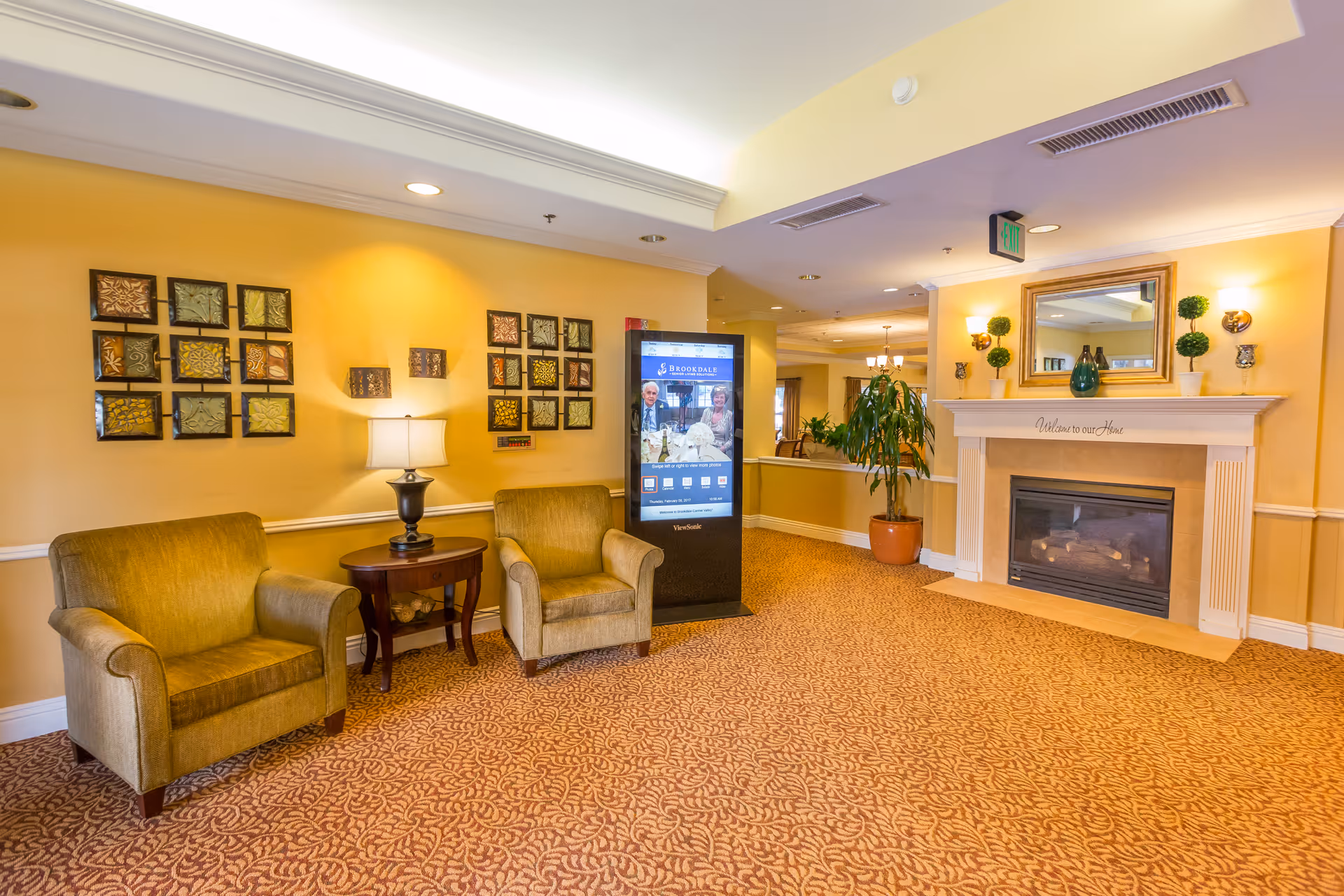 A cozy senior living facility common area with two beige armchairs separated by a small wooden table with a lamp. The walls are painted yellow and decorated with framed art pieces. There is a digital kiosk displaying community information, a fireplace with a mirror and decorative plants on the mantel, and a patterned carpet covering the floor.