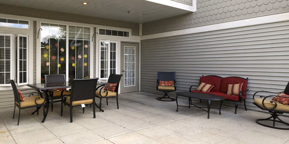 Covered outdoor patio with a dining table, chairs, and cushioned lounge seating along the building siding.