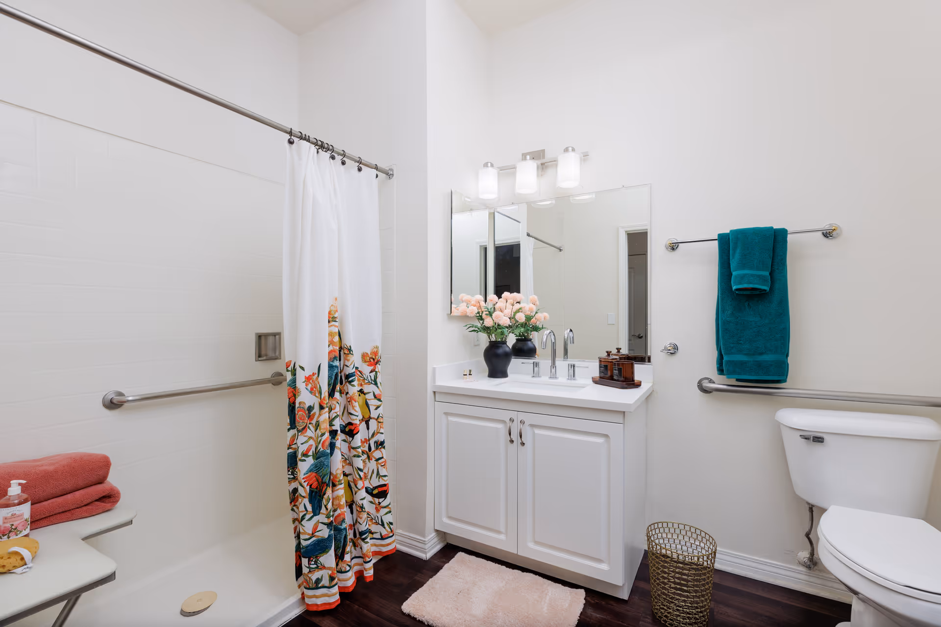 Bright bathroom with a floral shower curtain, white vanity and mirror with flowers, toilet, and teal towel on a rack.