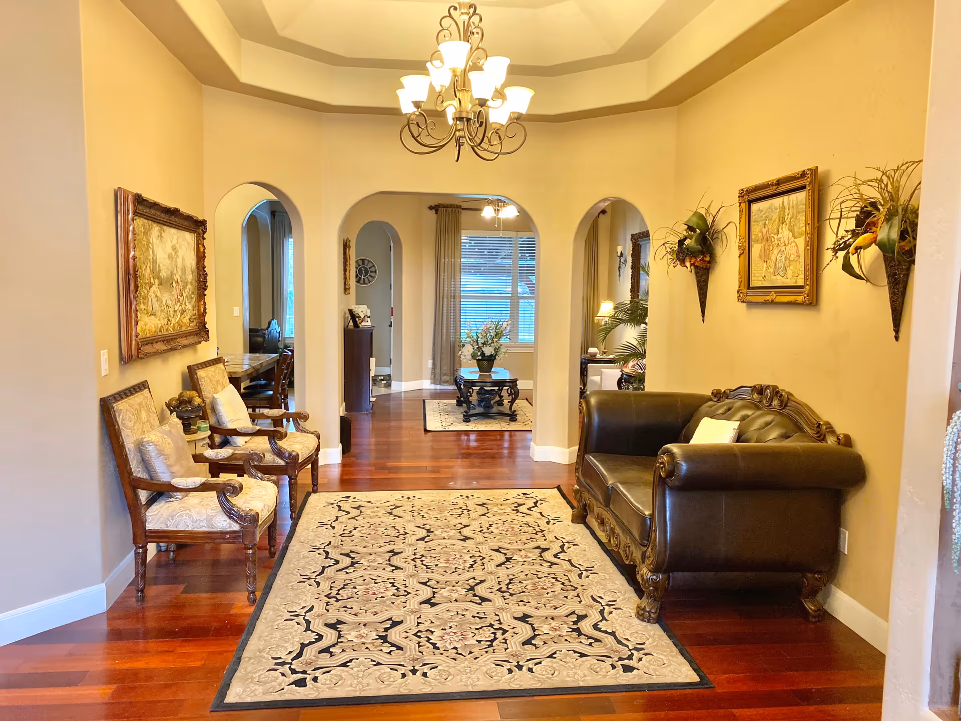 A warmly lit interior hallway with polished wooden floors and a decorative area rug. On the left side, there are two ornate wooden chairs with cushioned seats and a framed painting above them. On the right side, there is a dark leather loveseat with a pillow, a framed painting above it, and two wall-mounted floral arrangements. The hallway leads to an open area with arched doorways and a window with curtains in the background.