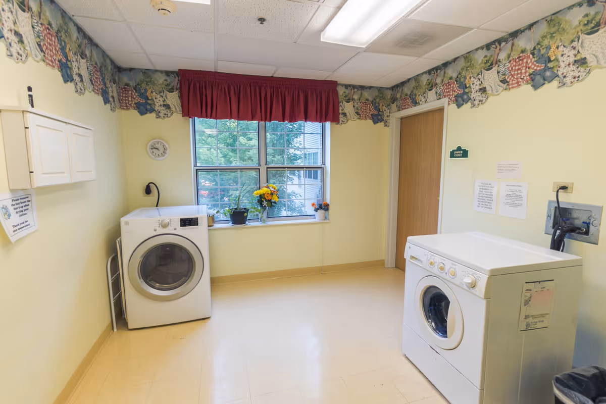 A small laundry room with a washer and dryer, a window with plants and a red valance, and pale yellow walls with floral border wallpaper.