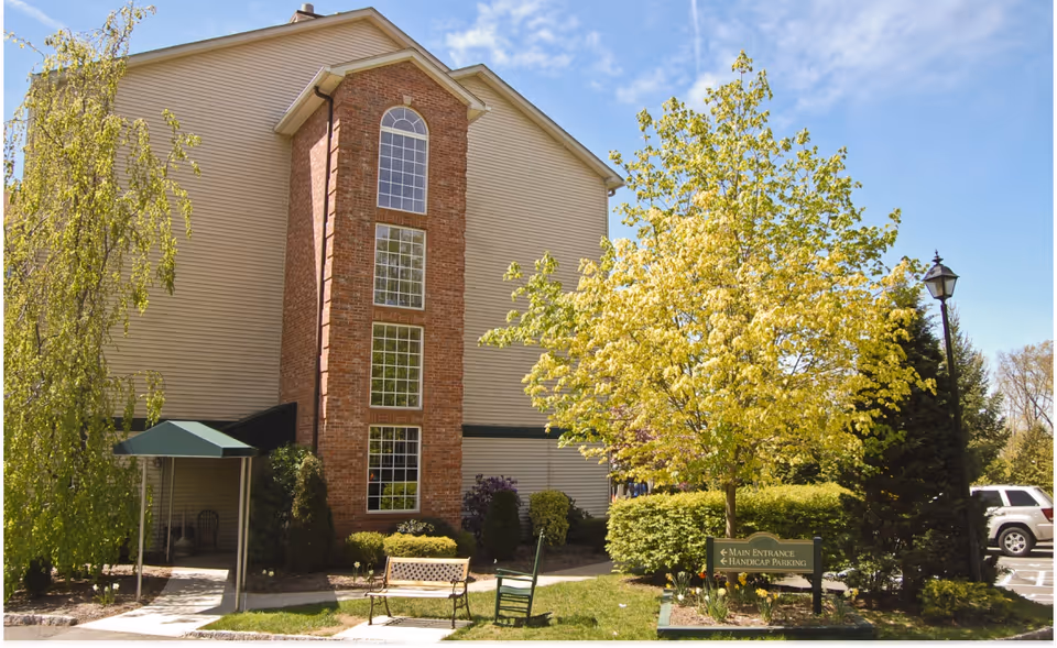Exterior view of a multi-story assisted living facility building with beige siding and a central brick section featuring tall windows. The entrance has a green awning, and the surrounding area includes green trees, bushes, a bench, a rocking chair, and a sign indicating the main entrance and handicap parking. A parking lot with cars is visible in the background under a blue sky with some clouds.