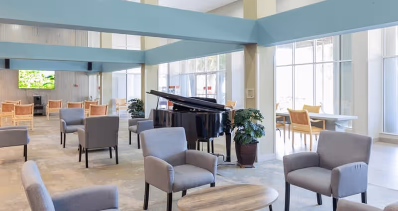 Bright senior living lounge with a grand piano, several armchairs and tables near large windows.