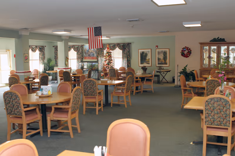 Dining room in a senior living facility with multiple round tables and upholstered chairs, a Christmas tree, and an American flag.