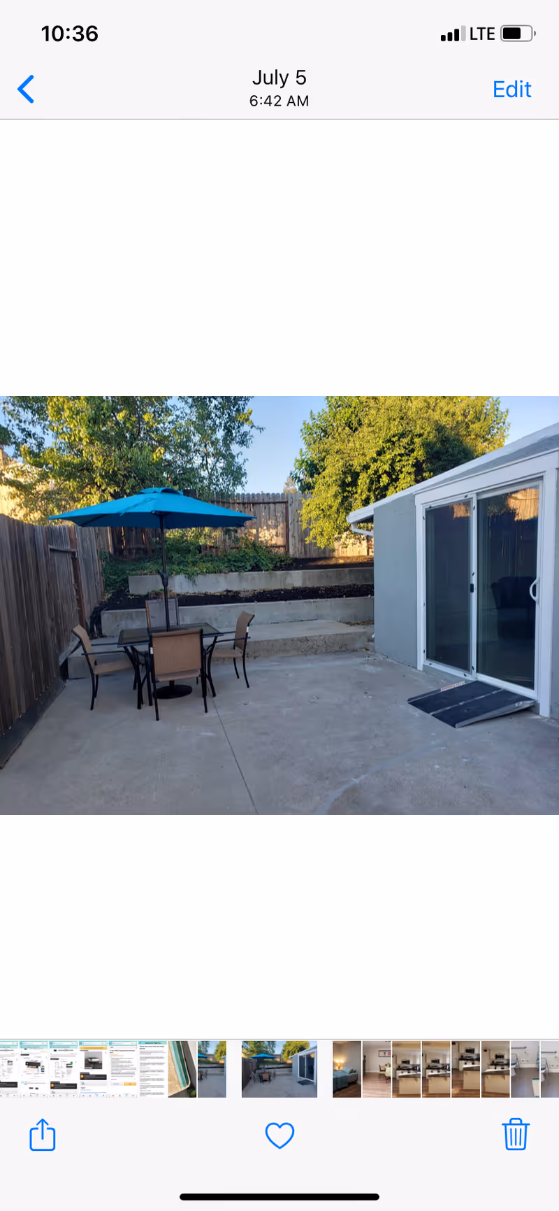 Outdoor patio area with a concrete floor, a table with four chairs, and a blue umbrella. There is a wooden fence on the left side and a gray building with sliding glass doors on the right. Trees and greenery are visible in the background.