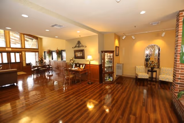Polished wood‑floored nursing home lobby with seating areas, a small table and lamp, display cabinet, and entrance doors.
