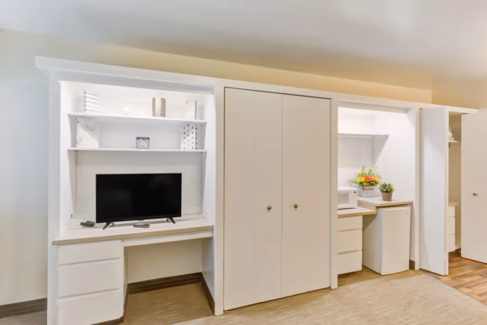 Built-in white cabinetry with a desk and flat-screen TV next to closets and a small kitchenette area with a microwave and mini fridge.