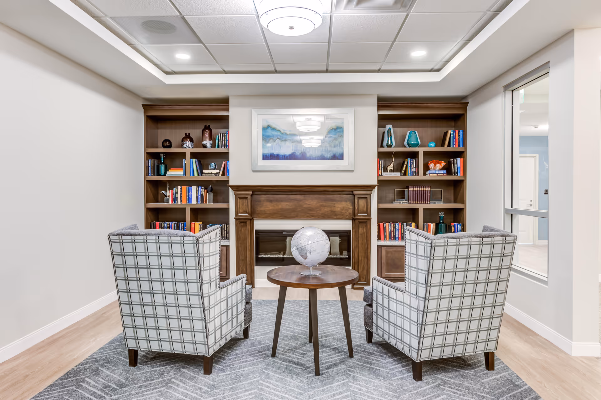 A cozy sitting area with two patterned armchairs facing a small round wooden table with a globe on it. Behind the table is a wooden fireplace flanked by two built-in bookshelves filled with books and decorative items. Above the fireplace hangs a framed abstract painting. The room has light-colored walls, a patterned rug, and a ceiling light fixture.