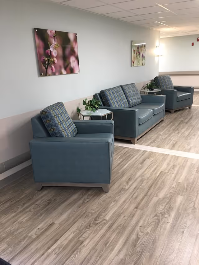 A hallway seating area in a senior living facility with three blue armchairs and one blue loveseat, each with patterned cushions. There are small side tables with green plants between the chairs. The walls are light-colored with two floral paintings hanging, and the floor has a wood-like finish.