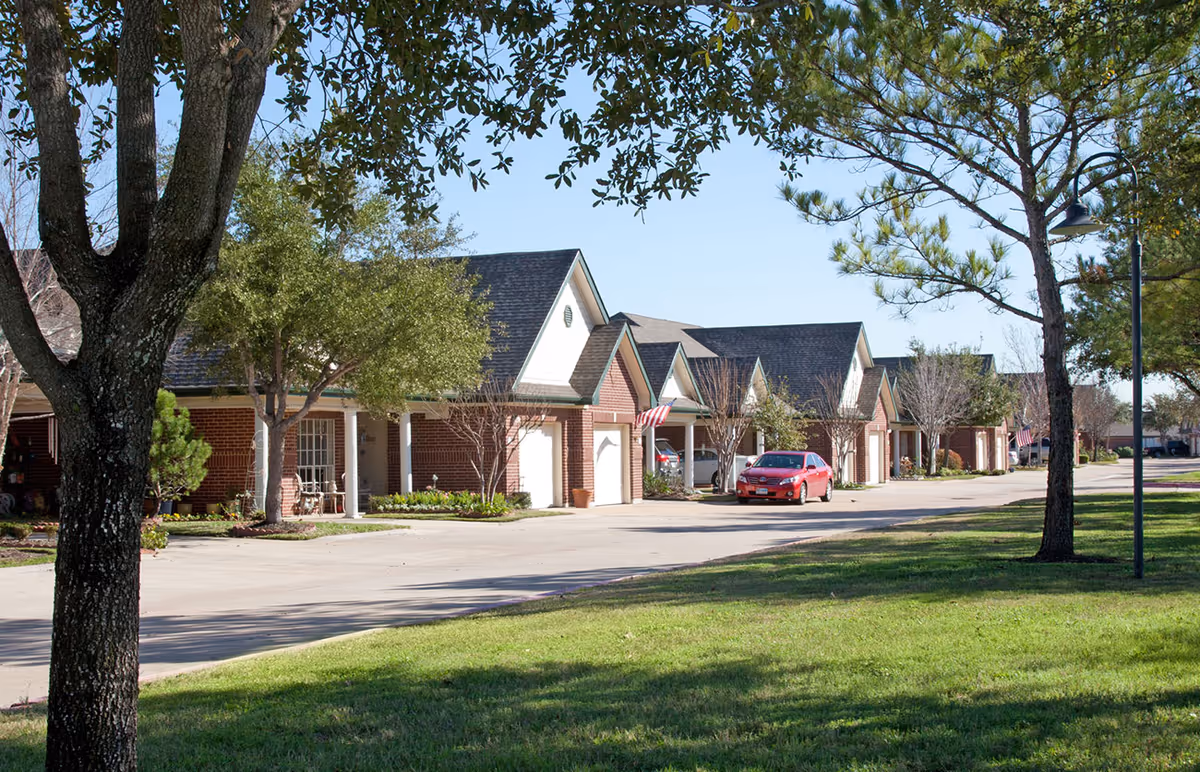A row of single-story brick residential buildings with garages, surrounded by trees and green grass under a clear blue sky. A red car is parked on the driveway in front of one of the garages.